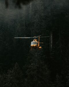 A yellow helicopter flying above a dense forest with tall evergreen trees.